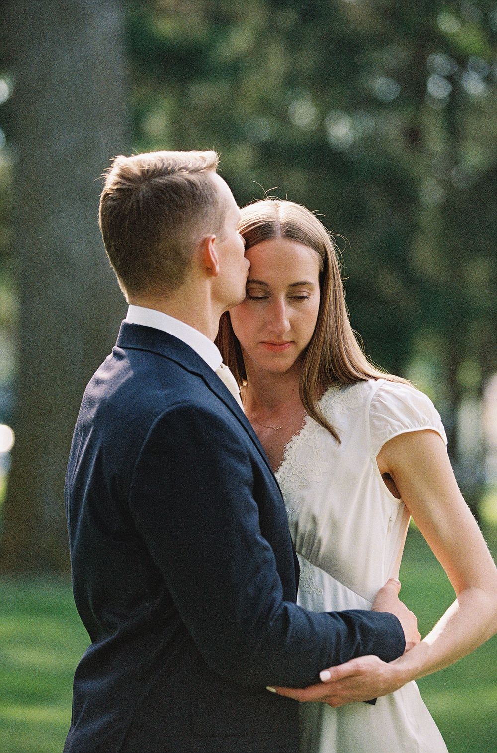 A groom in a navy suit kisses his bride on the forehead outdoors, as she closes her eyes in a white dress.
