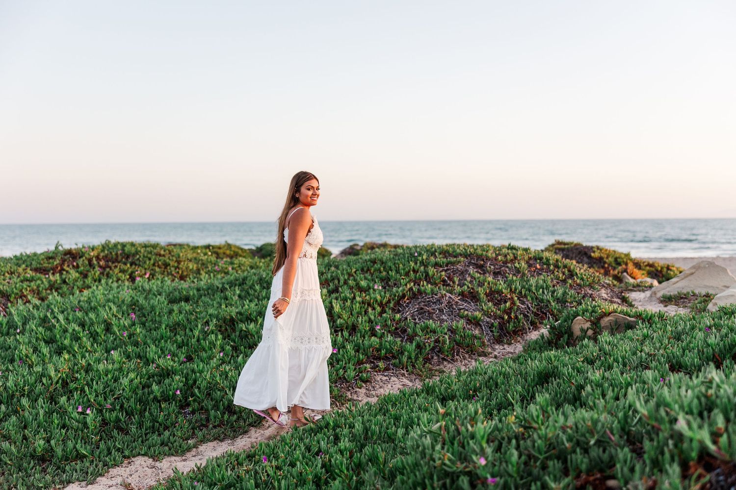 Woman in white maxi dress standing on coastal bluff with ocean view at sunset.