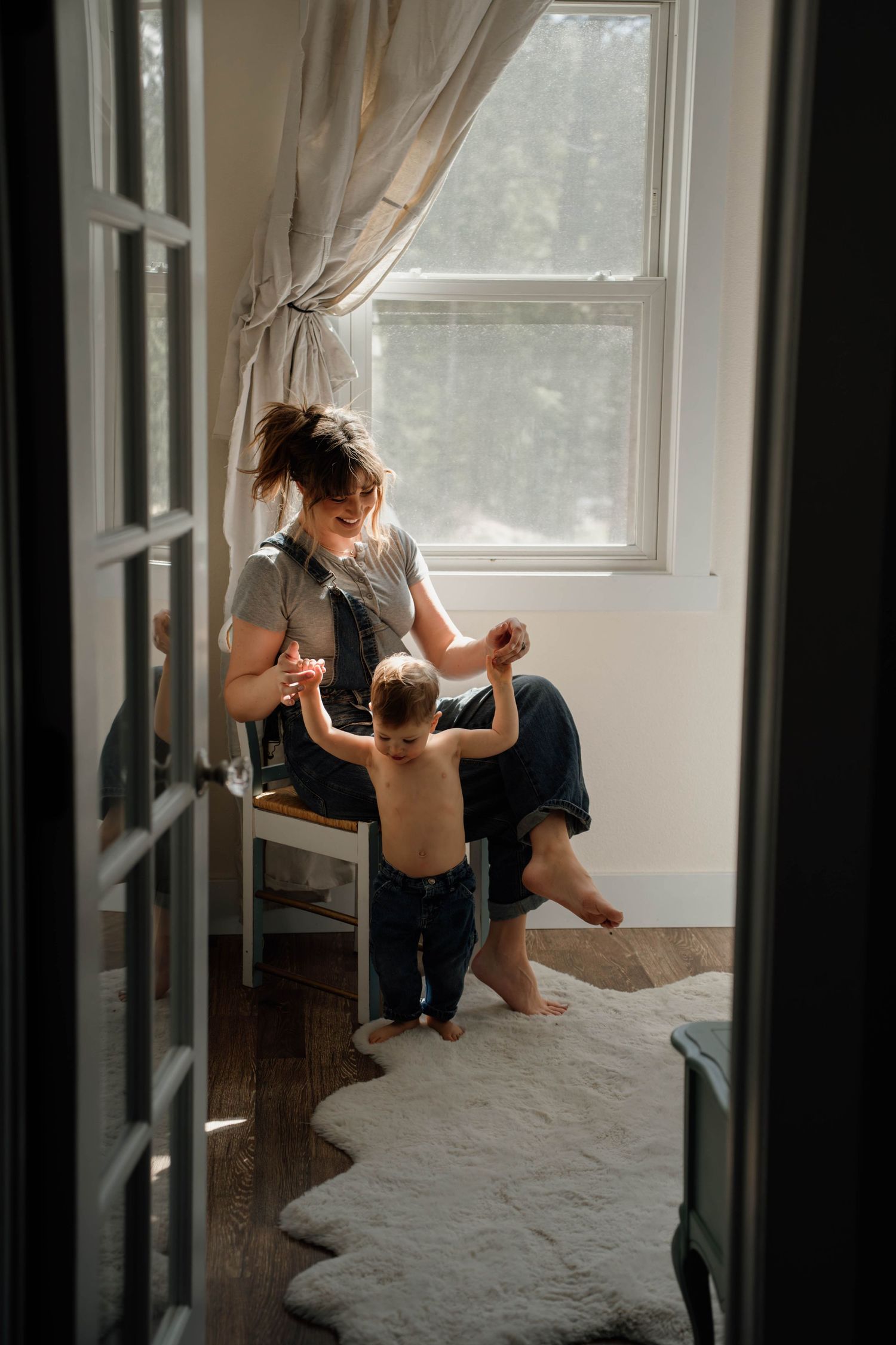 A mother sits by a window helping her toddler stand in a sunlit room with hardwood floors and a white rug.