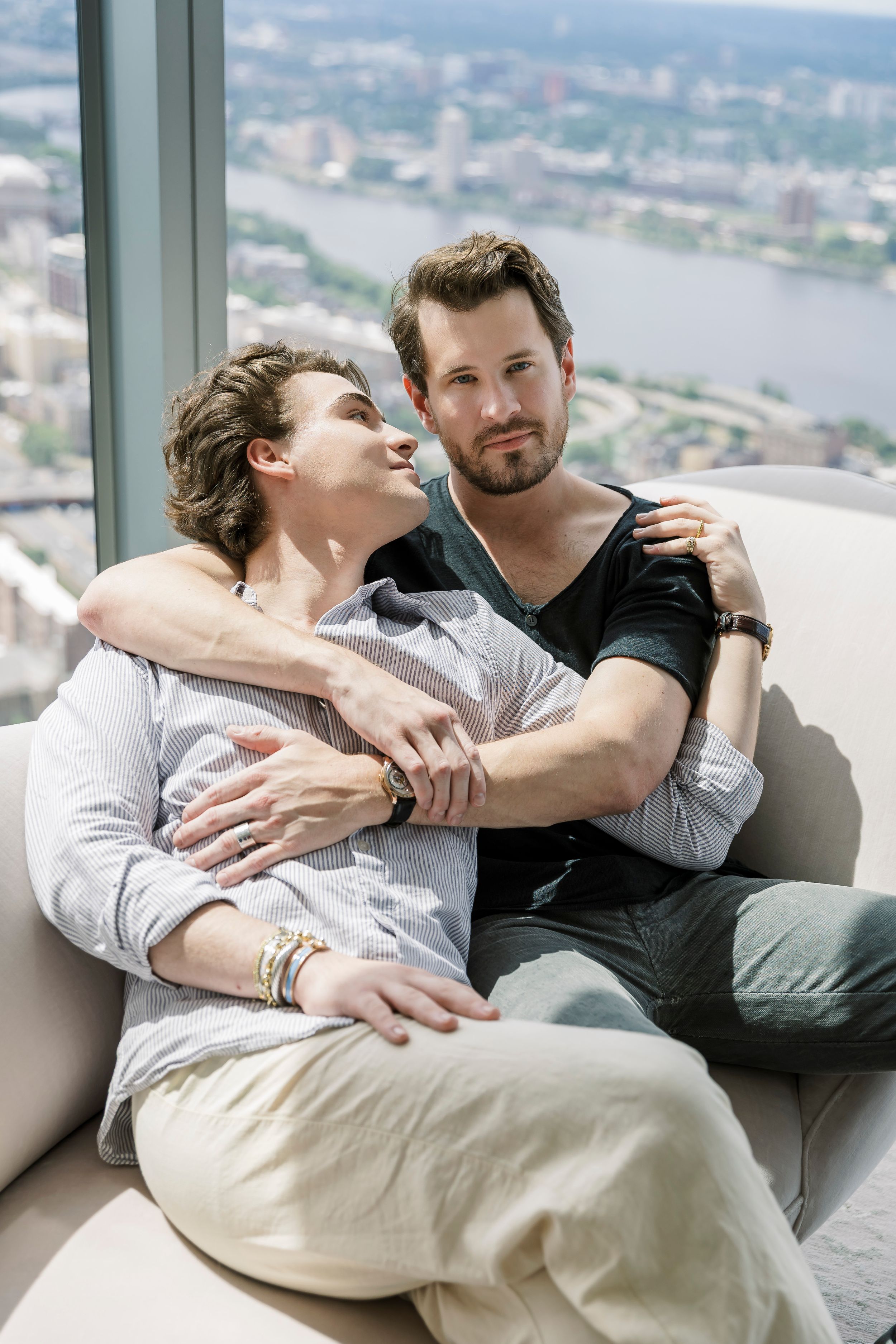 Couple lounging together on modern sofa with city skyline visible through window.