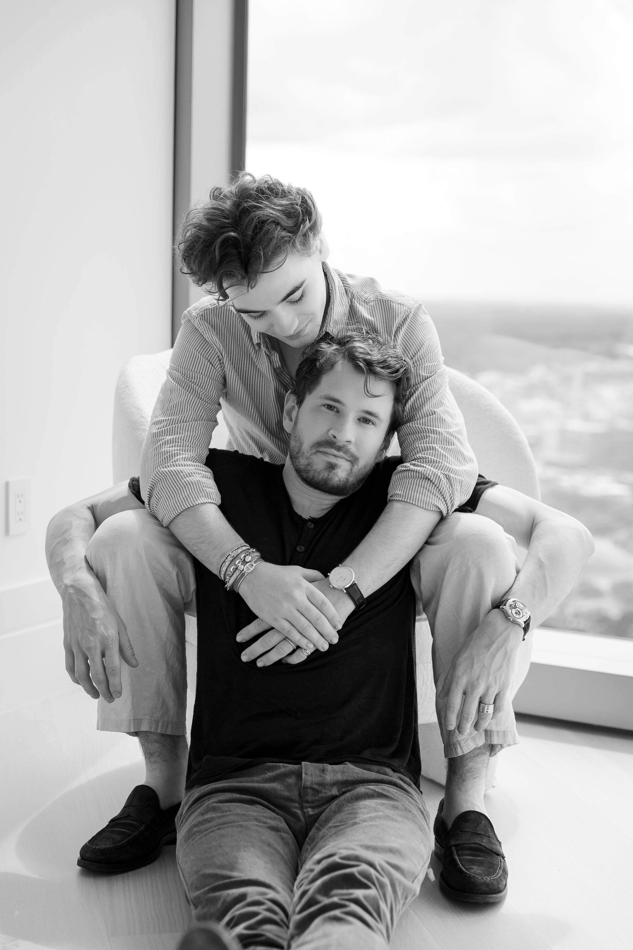 Two people sitting closely together on floor in front of large window with urban view.