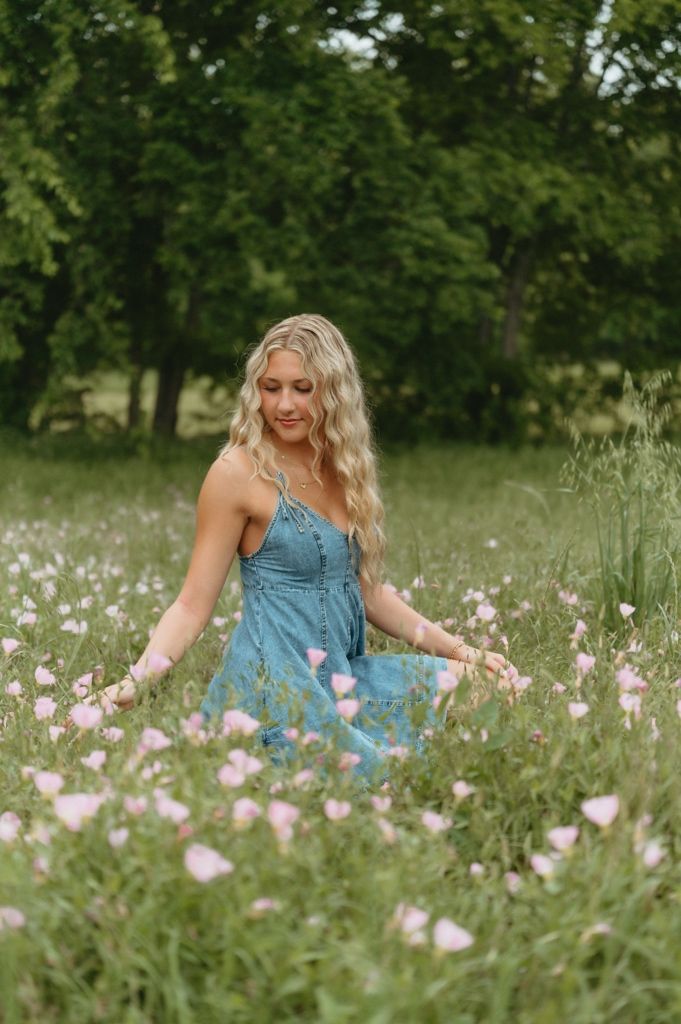 Blonde woman in blue dress touching pink wildflowers in a lush green meadow.