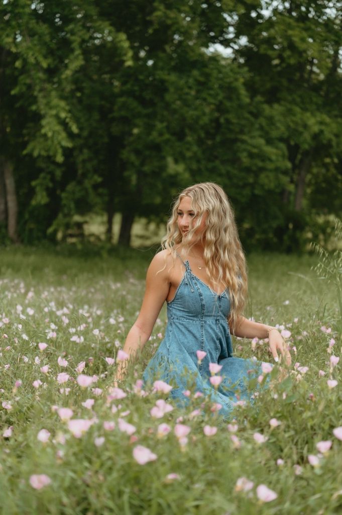 Blonde woman in blue denim dress sitting among pink wildflowers in a green meadow.
