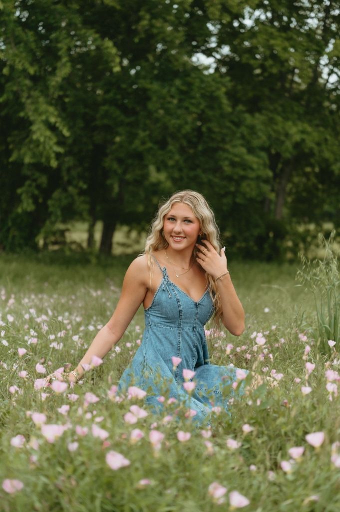 Young blonde woman in blue dress sitting in pink wildflower field with lush green trees in background.