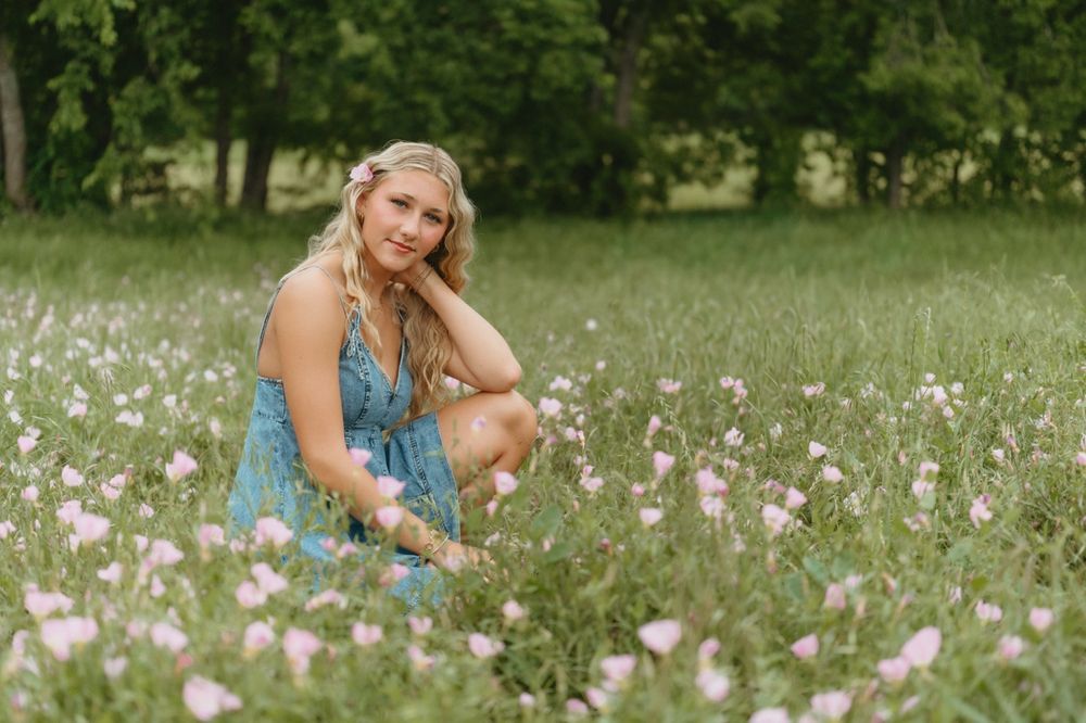 Young blonde woman in teal floral dress sitting in a field of pink wildflowers surrounded by lush green trees.