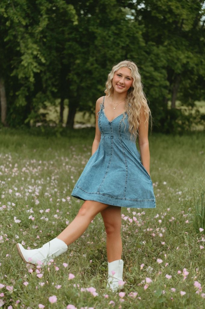 Happy blonde woman in denim dress and white boots standing in pink wildflower field.