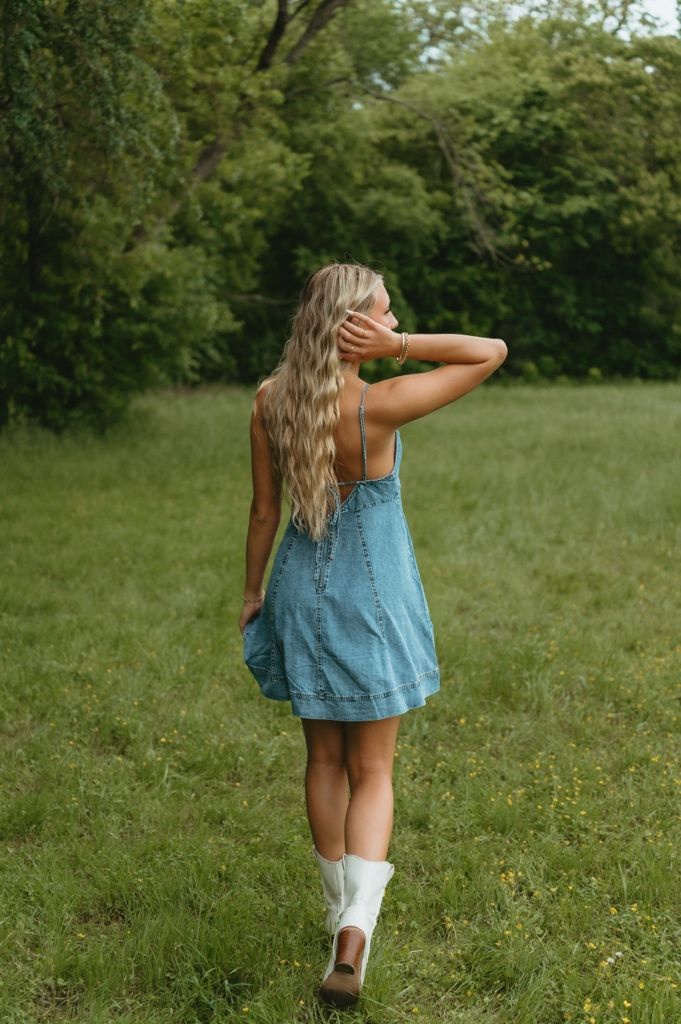 Blonde woman in denim dress and white boots walking away through lush green meadow.