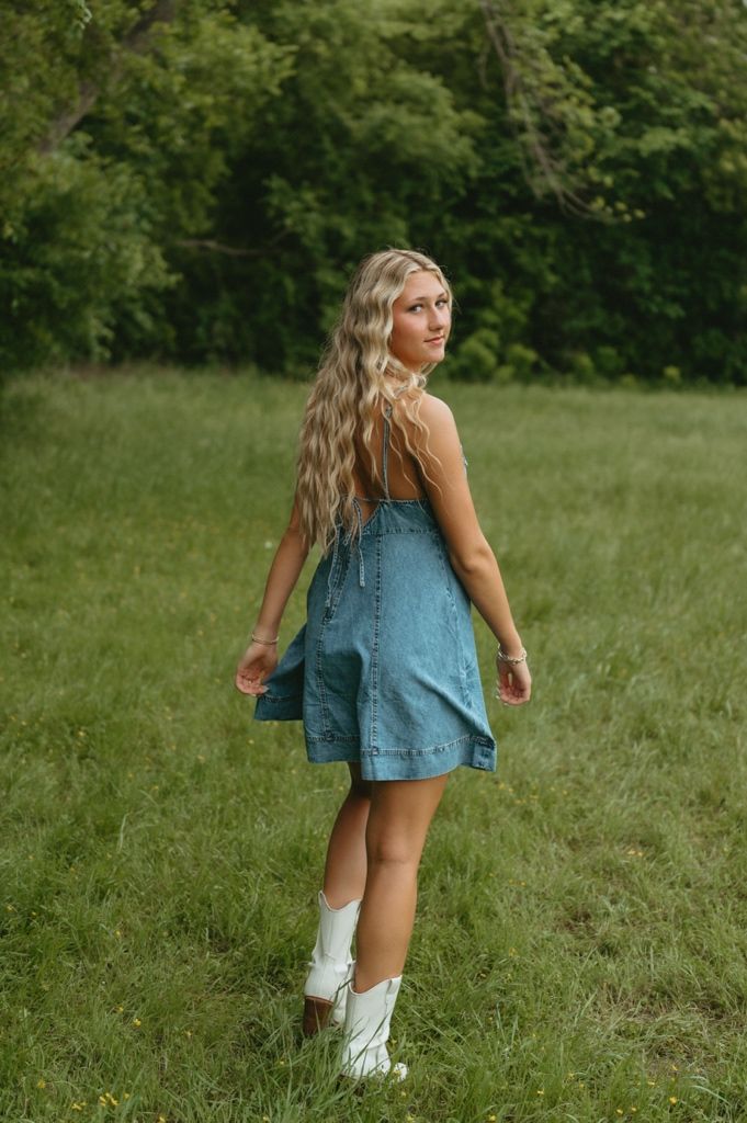 Blonde teen in denim mini dress and white cowboy boots twirling in green field during senior photo session.
