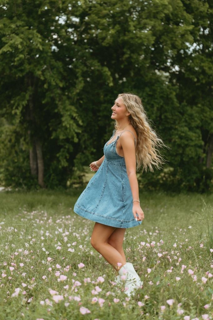 Young blonde woman in denim dress twirling in a wildflower meadow with lush green trees in the background.