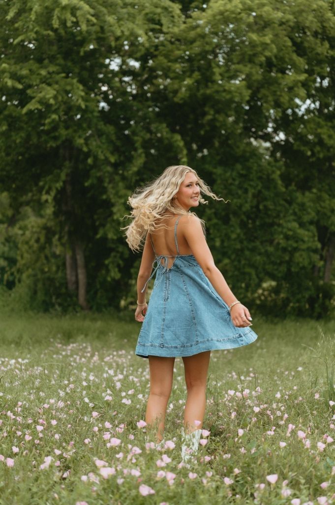 Blonde woman twirling in denim mini dress among wildflowers in green meadow, hair flowing, wearing white boots.