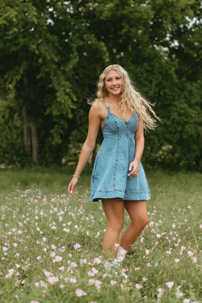 Smiling blonde woman in denim sundress walking through pink wildflower meadow with green trees background.