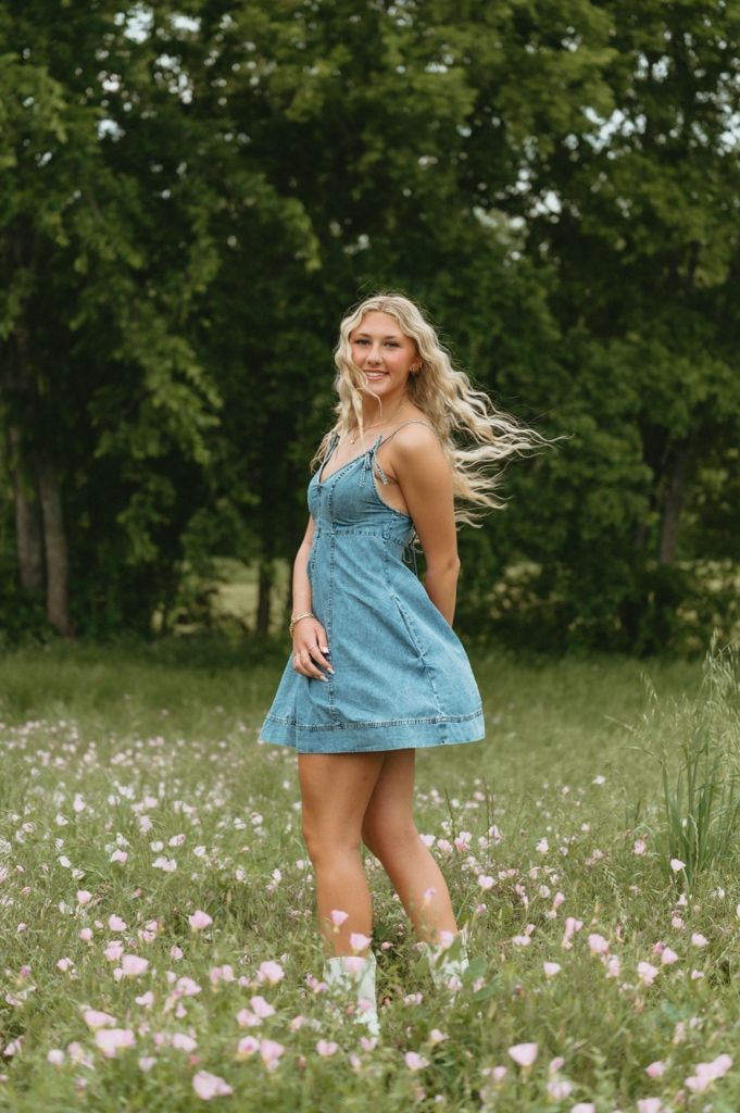 Young blonde woman in denim dress and white boots smiling in a wildflower meadow with green trees behind her.