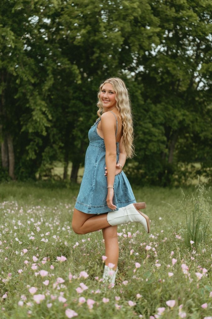 Smiling blonde in denim dress and white cowboy boots posing in pink wildflower meadow.