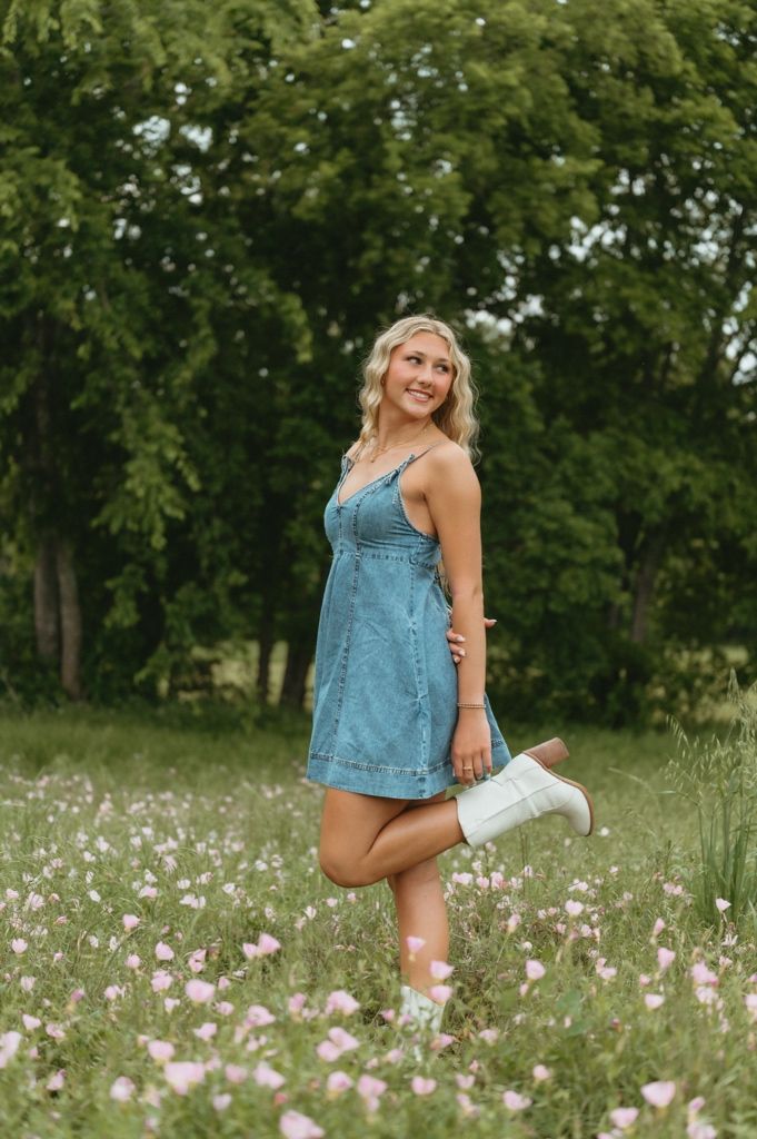 Blonde woman in blue denim dress and white cowboy boots posing playfully in a blooming wildflower field.