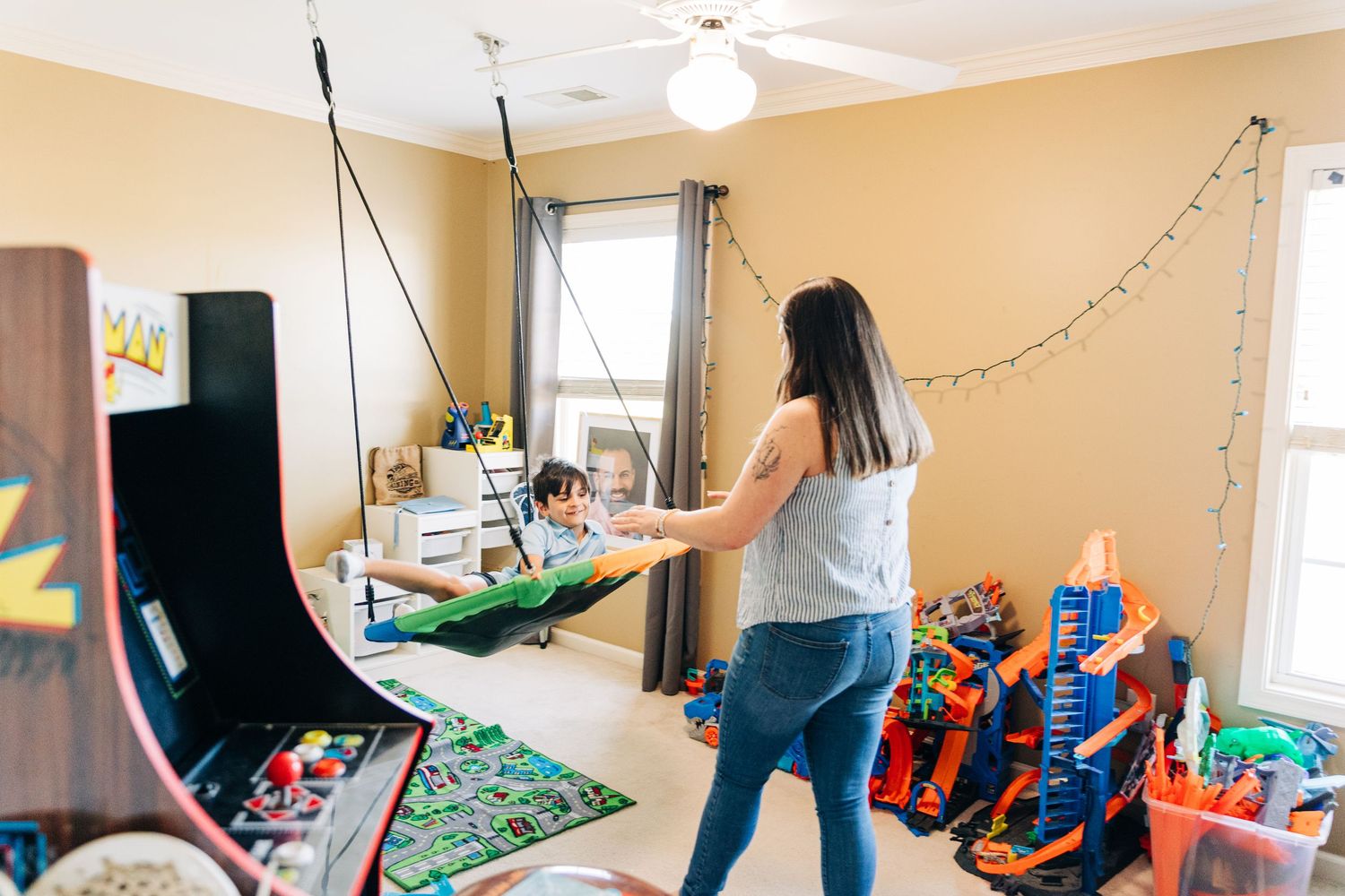 Mother pushing child in indoor hammock swing in colorful playroom filled with toys and arcade games.