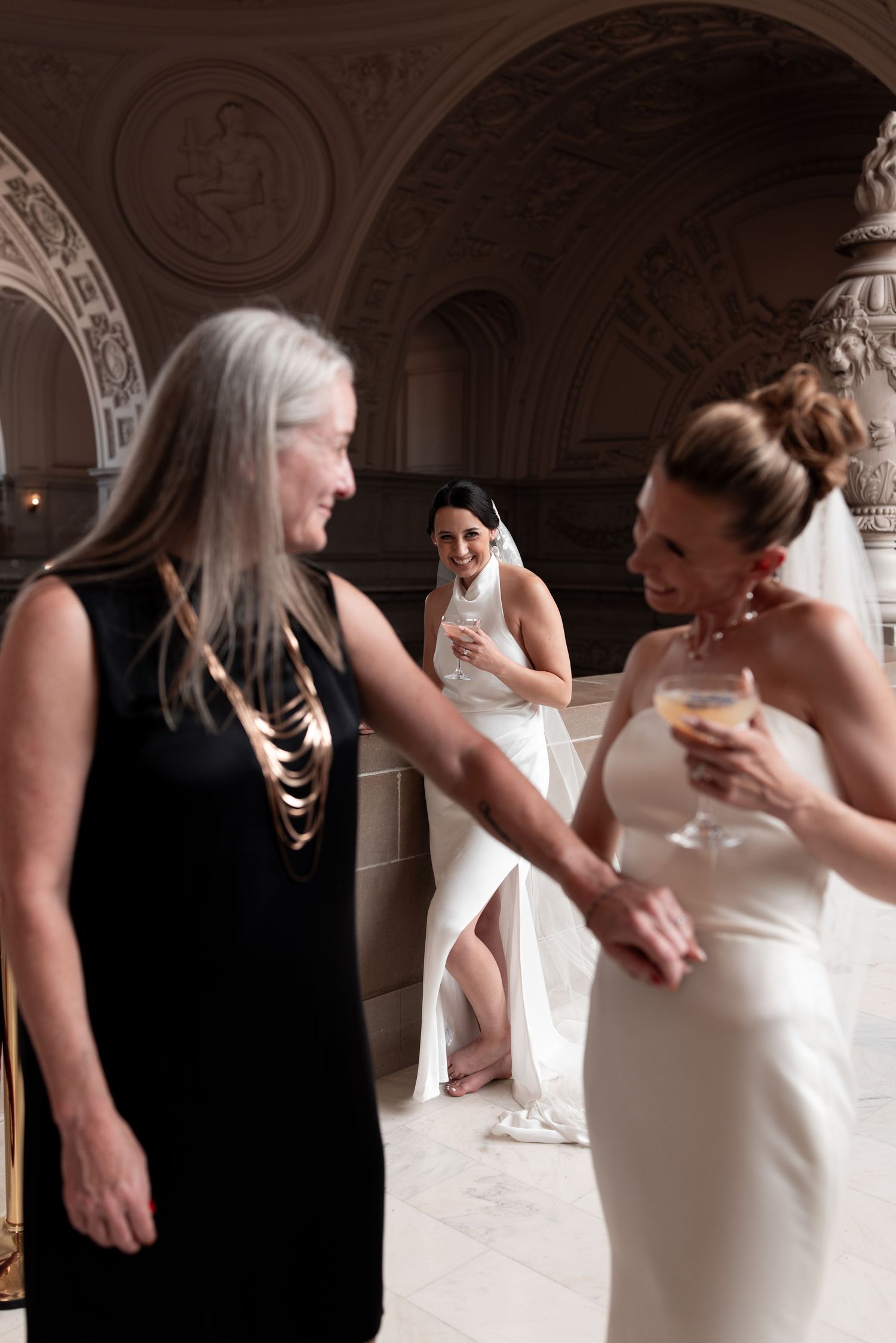 Two brides in white gowns celebrate with champagne alongside a guest in a black dress in an ornate wedding venue.