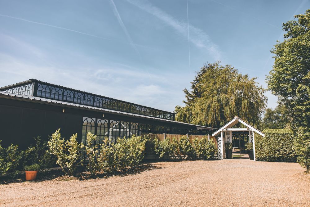 A charming greenhouse venue with lush hedges, gravel driveway, and a covered entrance under a clear blue sky.