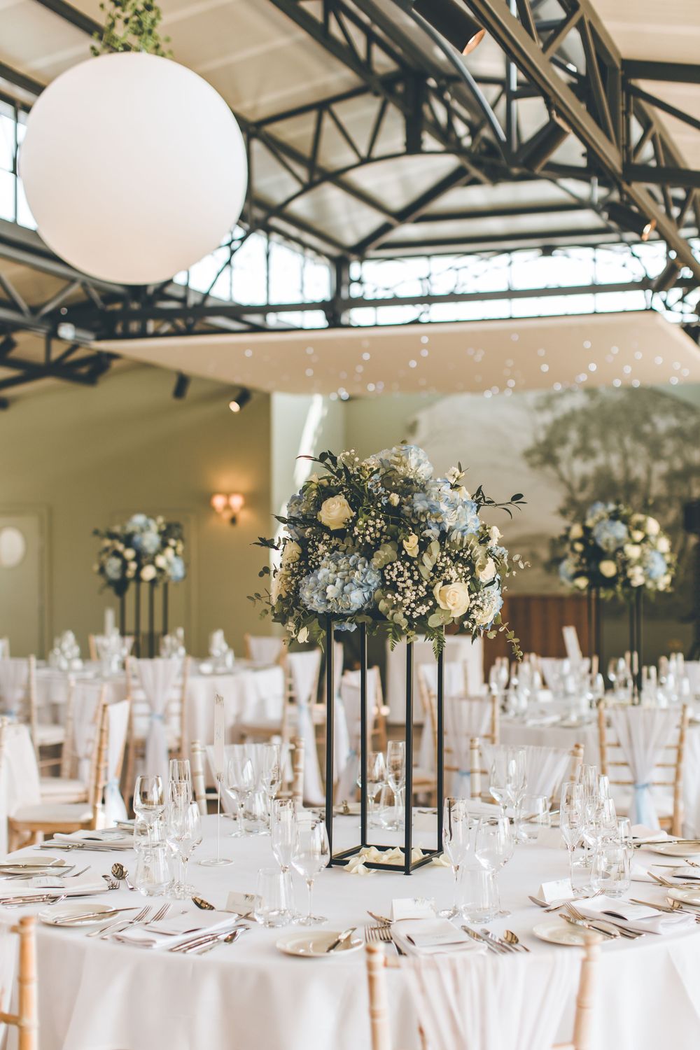 Elegant wedding reception hall with tall floral centerpieces, white balloon decor, and glass ceiling.