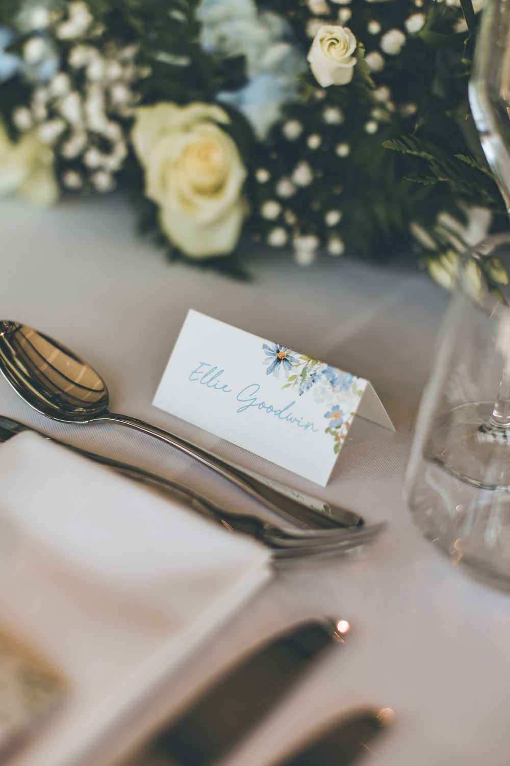 Wedding place card reading Ellie Goodwin beside white roses and baby's breath centerpiece.