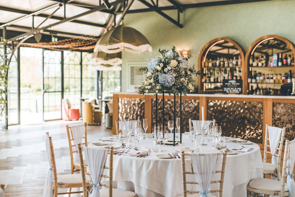 Bright wedding reception venue with tall floral centerpieces, gold chairs, and a bar backdrop.