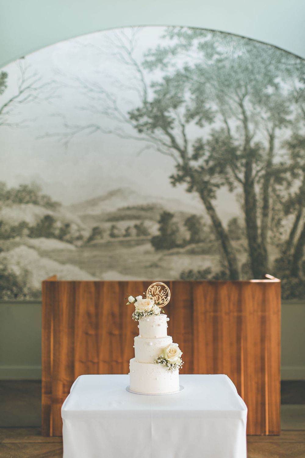 Three-tier white wedding cake with floral decorations on a white table before a scenic mural.