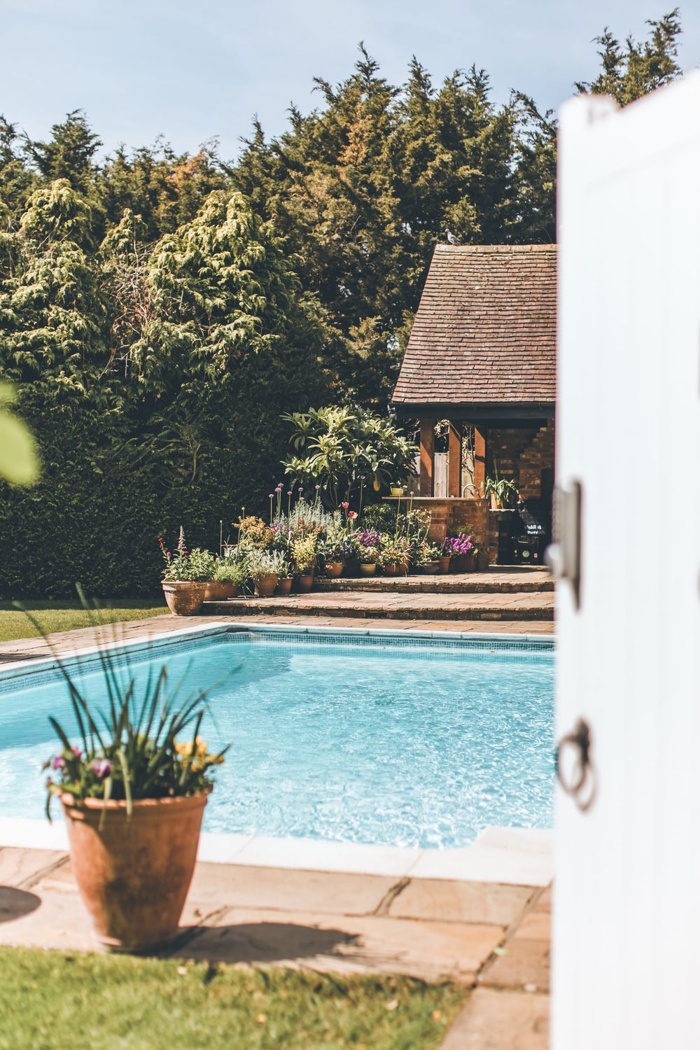 Sunny garden with a rectangular swimming pool, potted plants, and a stone pool house.