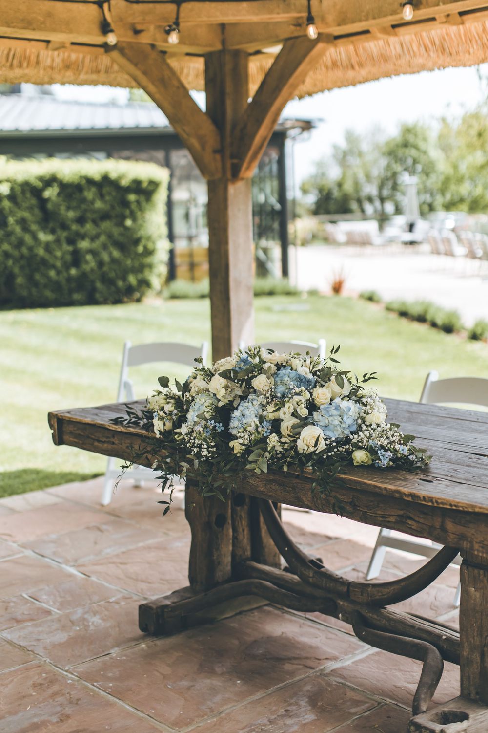Rustic wooden ceremony table adorned with blue hydrangea and white rose floral arrangement under pergola.