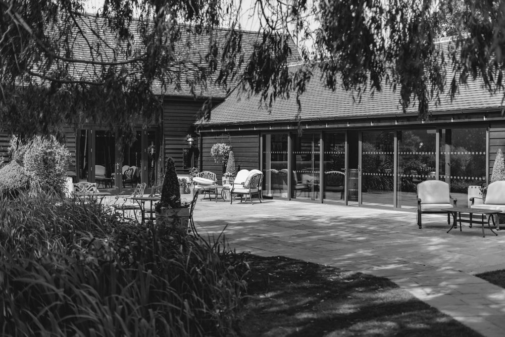 Black and white photo of a rustic barn wedding venue with outdoor patio seating surrounded by lush trees.