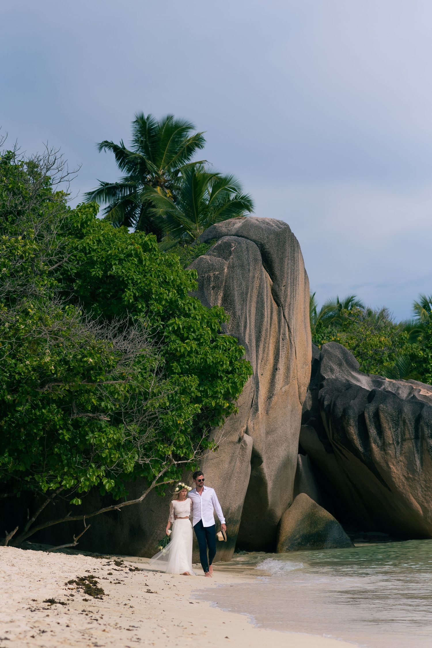 Couple walking hand in hand on a tropical beach with large granite boulders and lush palm trees in the Seychelles.