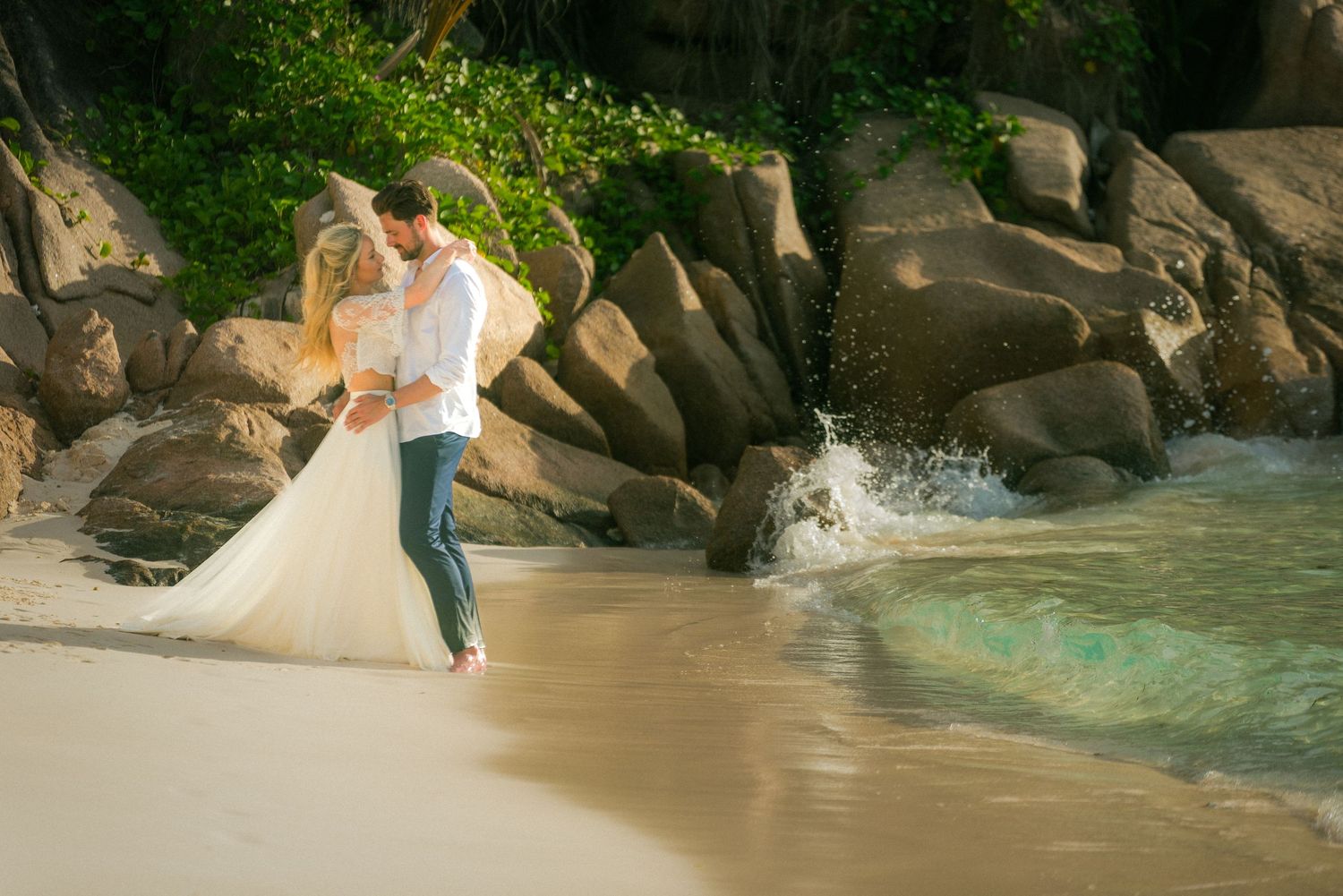 Bride and groom embracing on a tropical beach with turquoise waves and large granite rocks in the background.