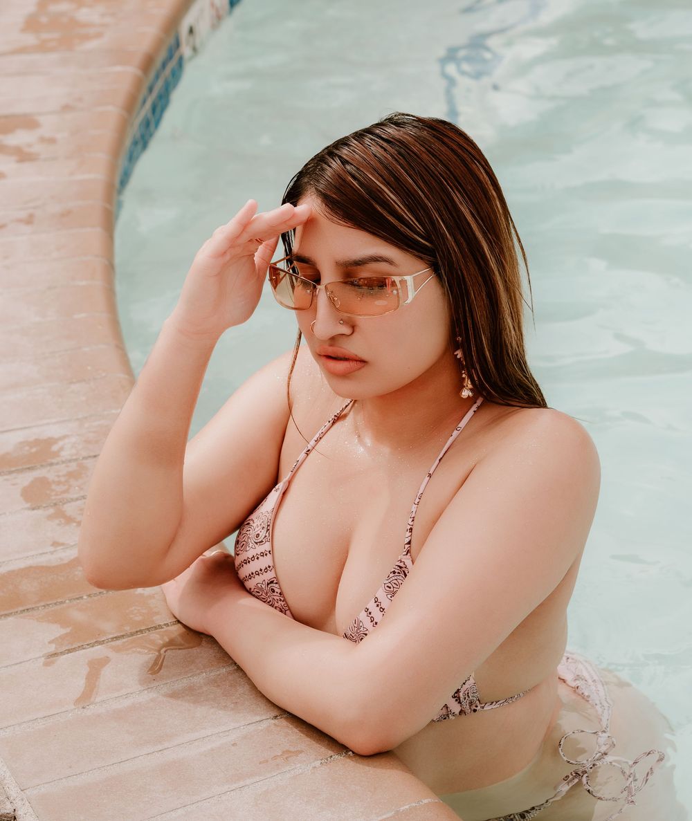 Young woman in patterned bikini and sunglasses relaxing at pool edge on sunny day.