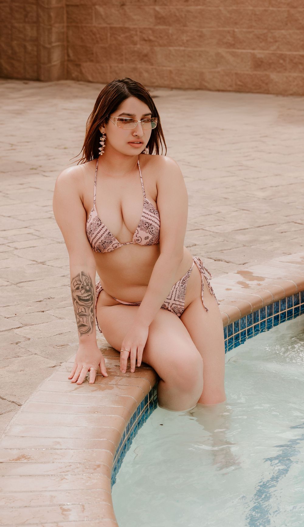 Woman in patterned bikini with tattoo posing by pool edge against stone wall backdrop.