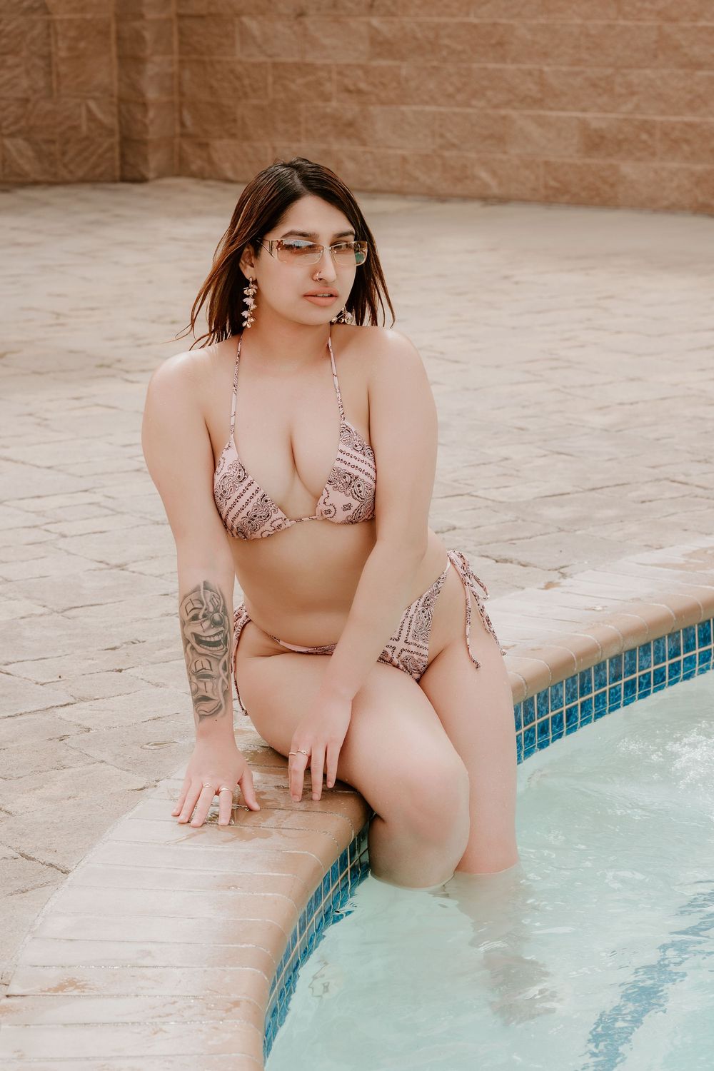 Woman in patterned bikini with tattoo sitting poolside, wearing sunglasses, by tiled pool.