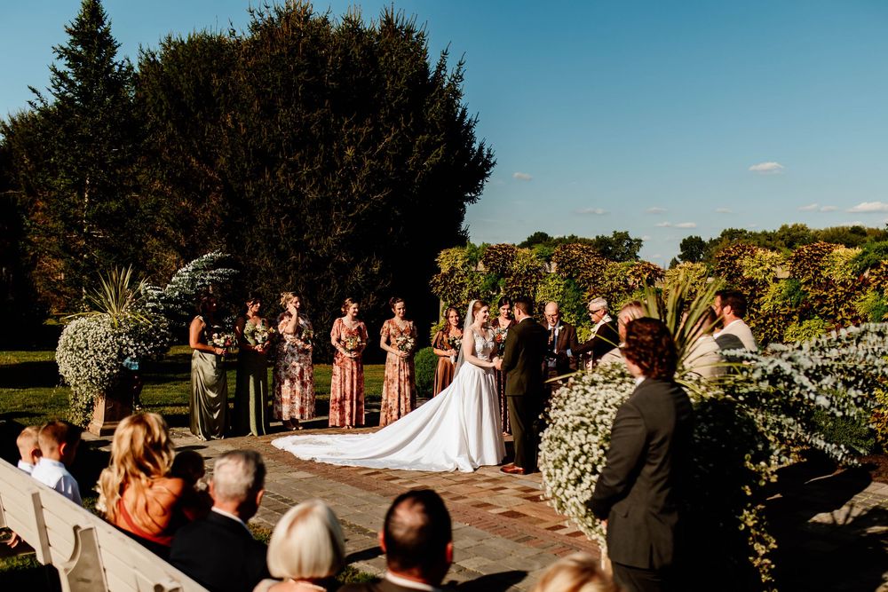 Outdoor wedding ceremony with bride and groom at altar, surrounded by bridesmaids and groomsmen, lush greenery under blue sky.