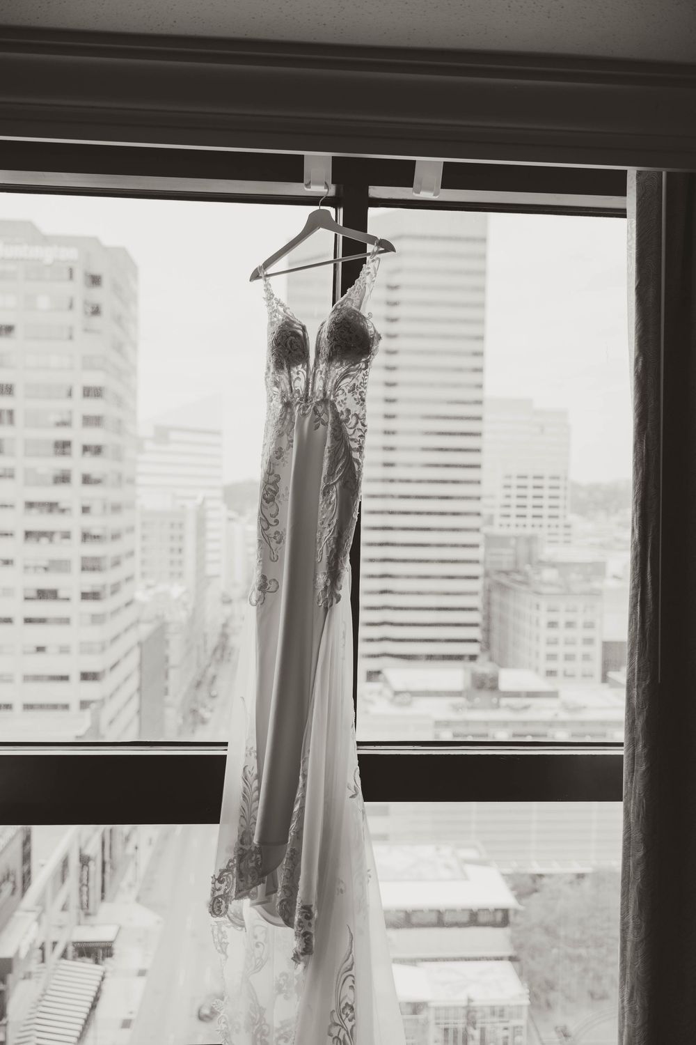 Black and white photo of an elegant lace wedding gown hanging in a hotel window with city skyline views.