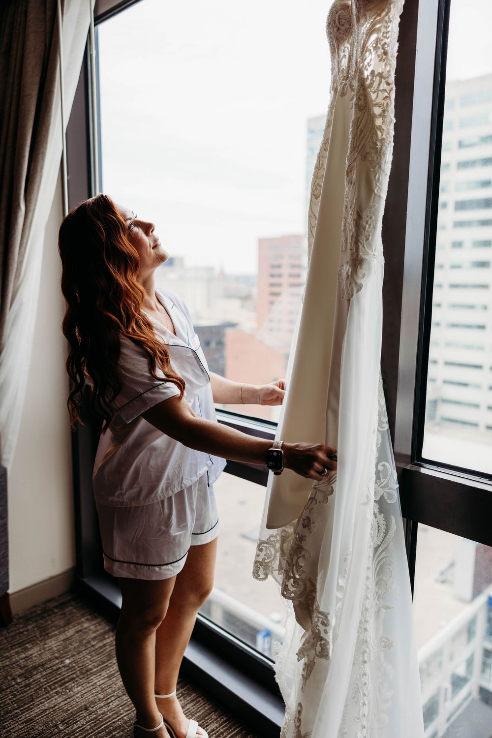 Bride in pajamas admiring her lace wedding dress hanging by a hotel window with city views.