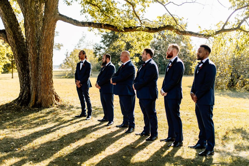 Groomsmen in navy suits standing under a large oak tree at an outdoor wedding venue on a sunny day.