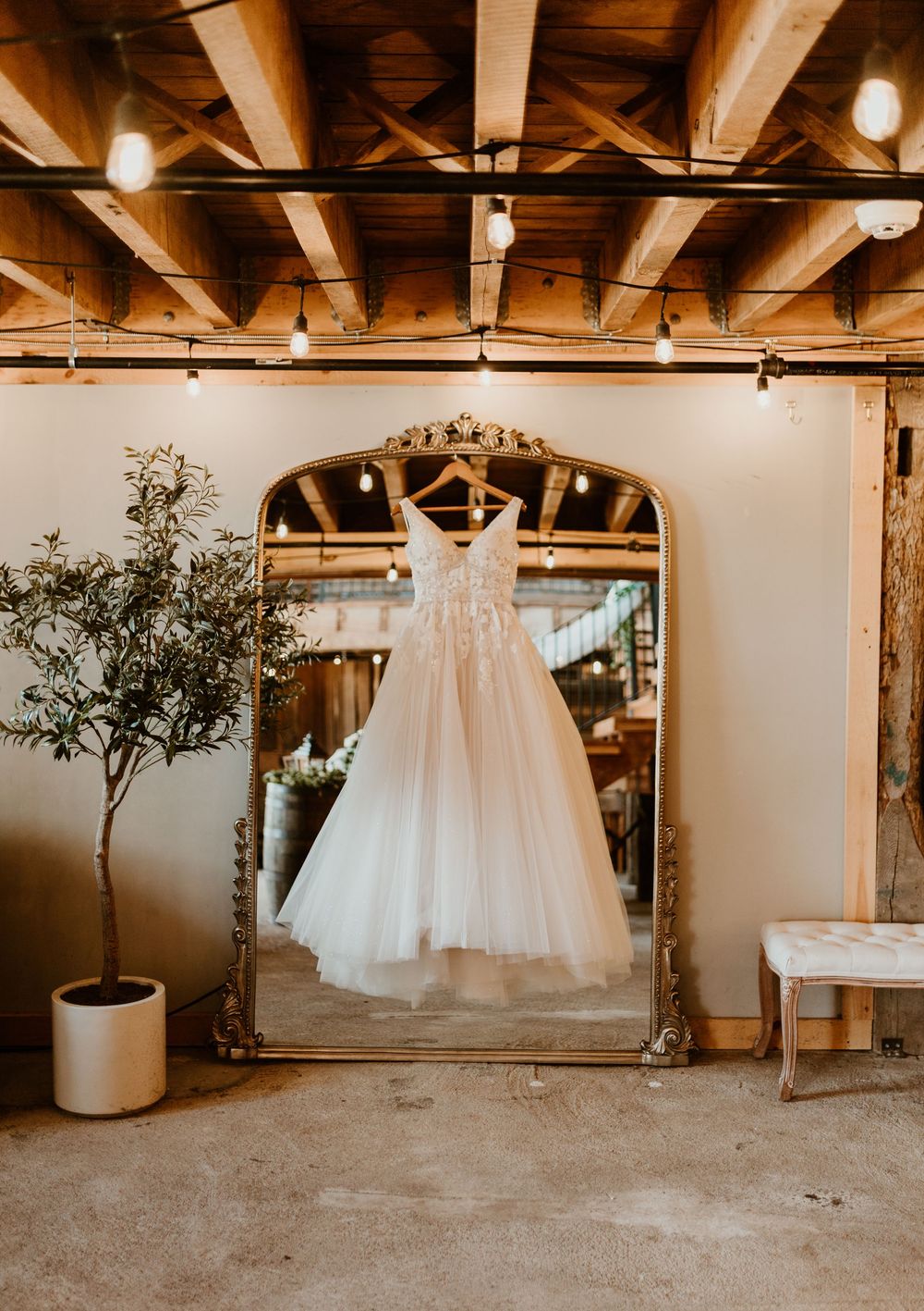 A flowing white wedding dress hangs on a ornate gold mirror in a rustic barn venue with string lights.