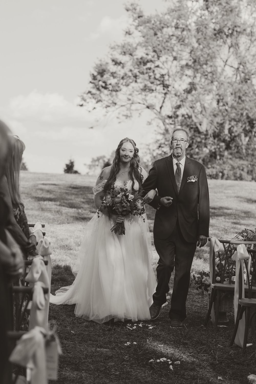 Black and white photo of a bride walking down the aisle with an older gentleman at an outdoor wedding ceremony.