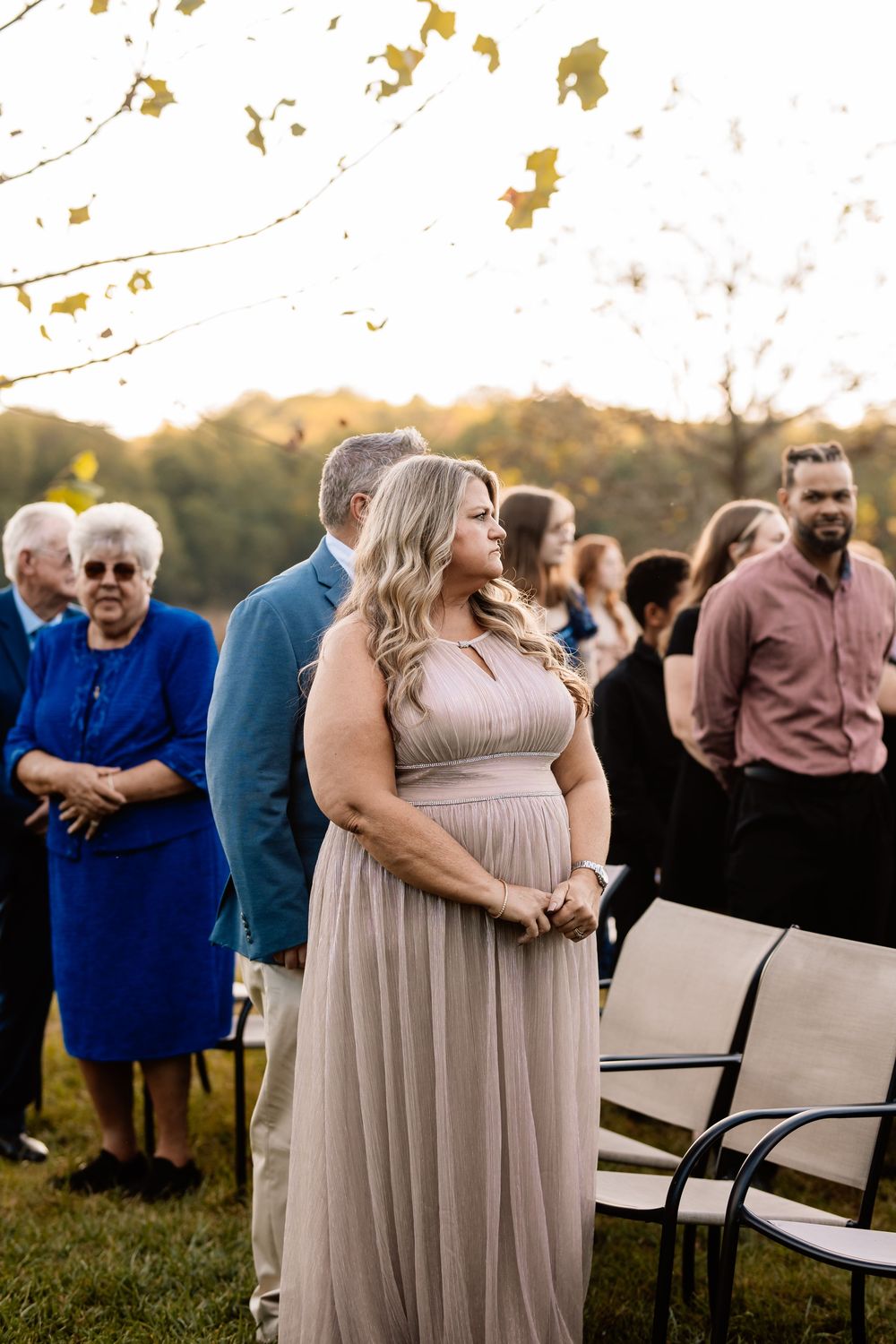 A woman in a taupe gown stands at an outdoor wedding ceremony at golden hour, surrounded by guests.