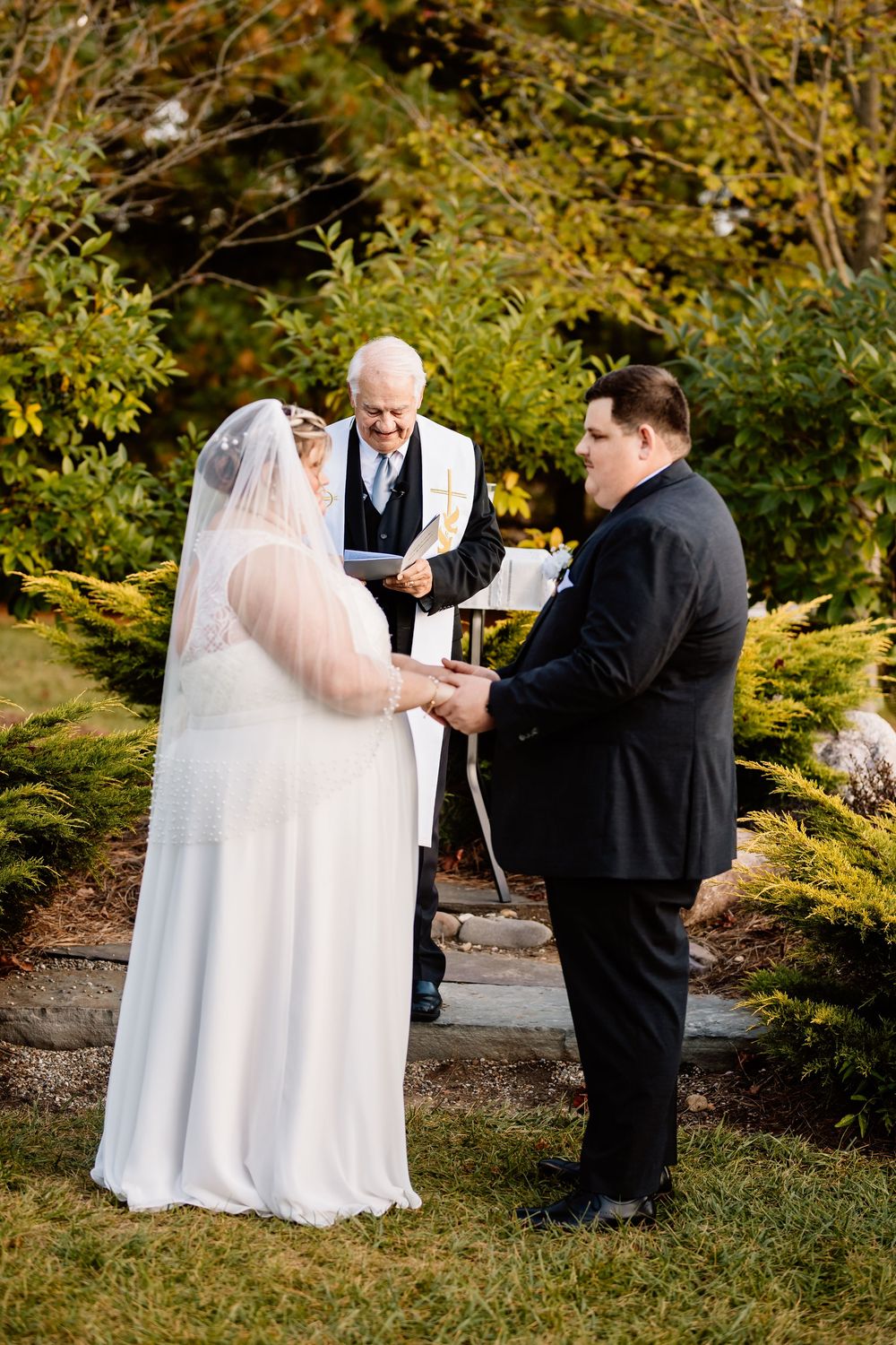 Outdoor wedding ceremony with bride in white veil, groom in black suit, and officiant reading vows in a garden setting.