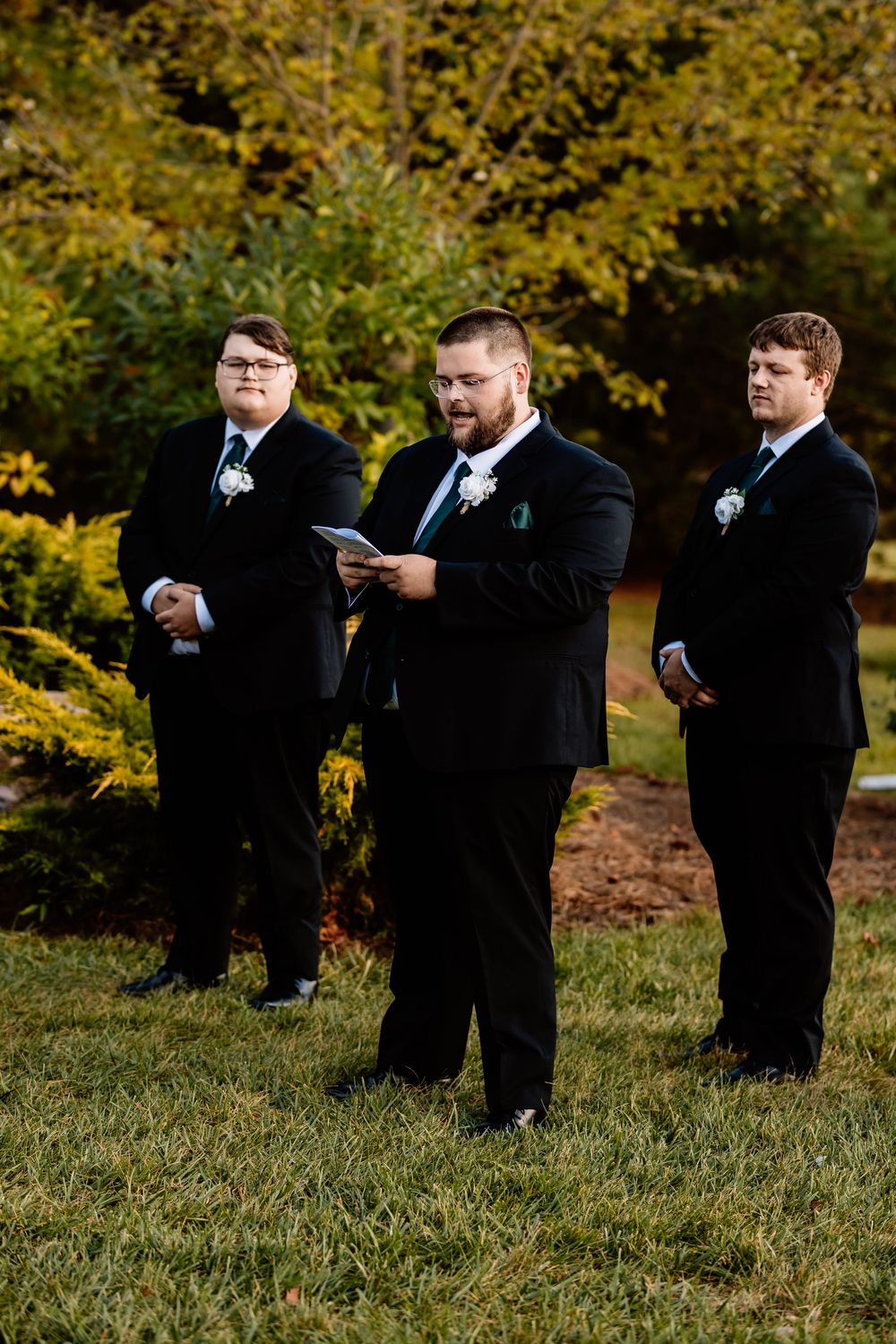 Three groomsmen in black suits with boutonnieres stand outdoors during a wedding ceremony amid autumn foliage.