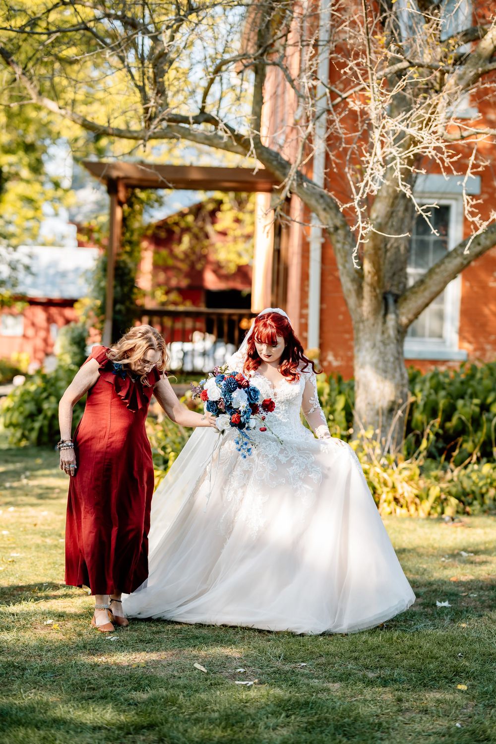 Bride in white gown holds bouquet while bridesmaid in red dress assists her walking outdoors near brick building.