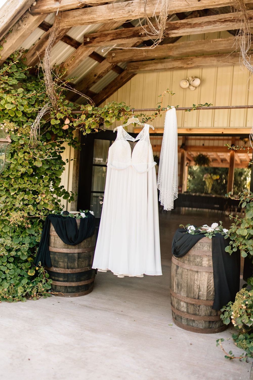 White wedding dress hanging at rustic barn entrance with wooden barrels and green ivy decor.