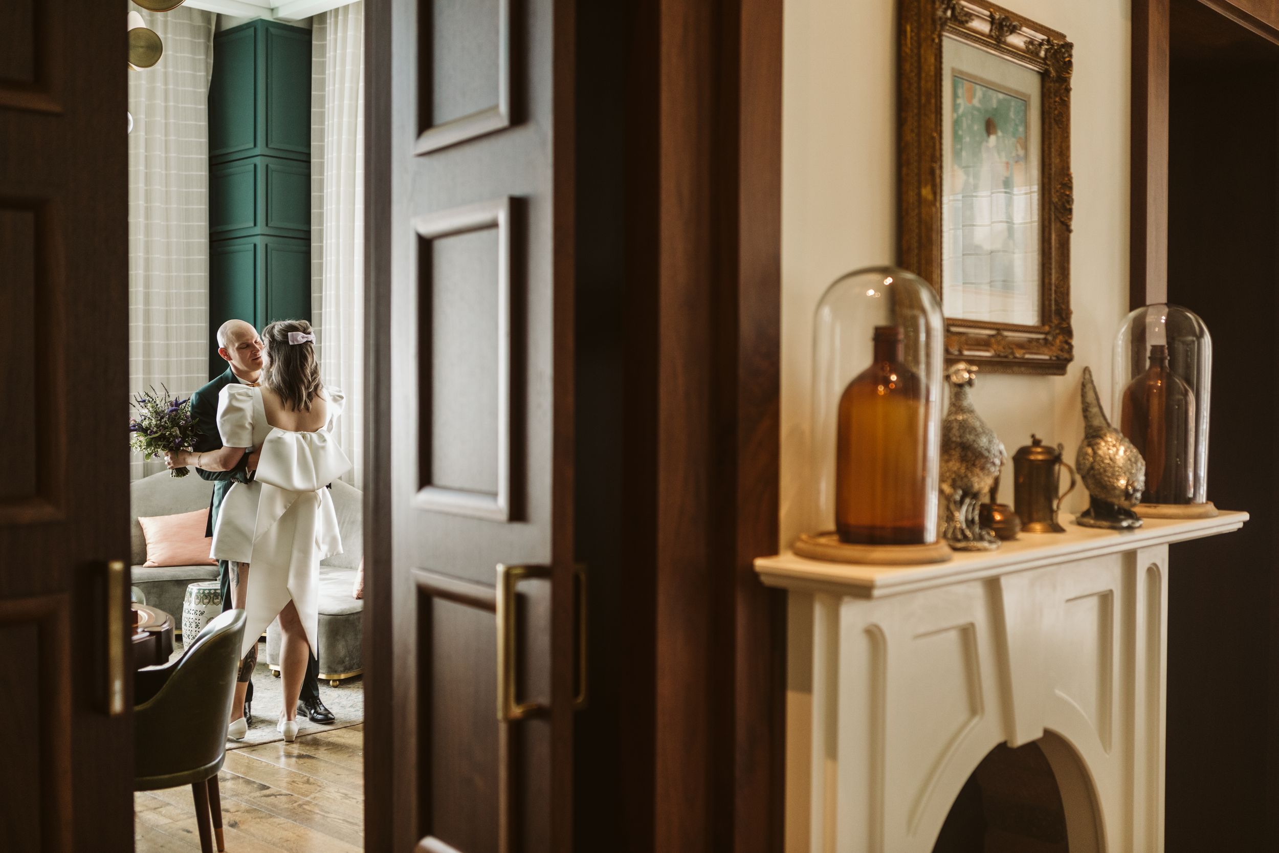 Elegant interior hallway with dark wood door, white fireplace mantel with glass cloches, and couple visible in background.