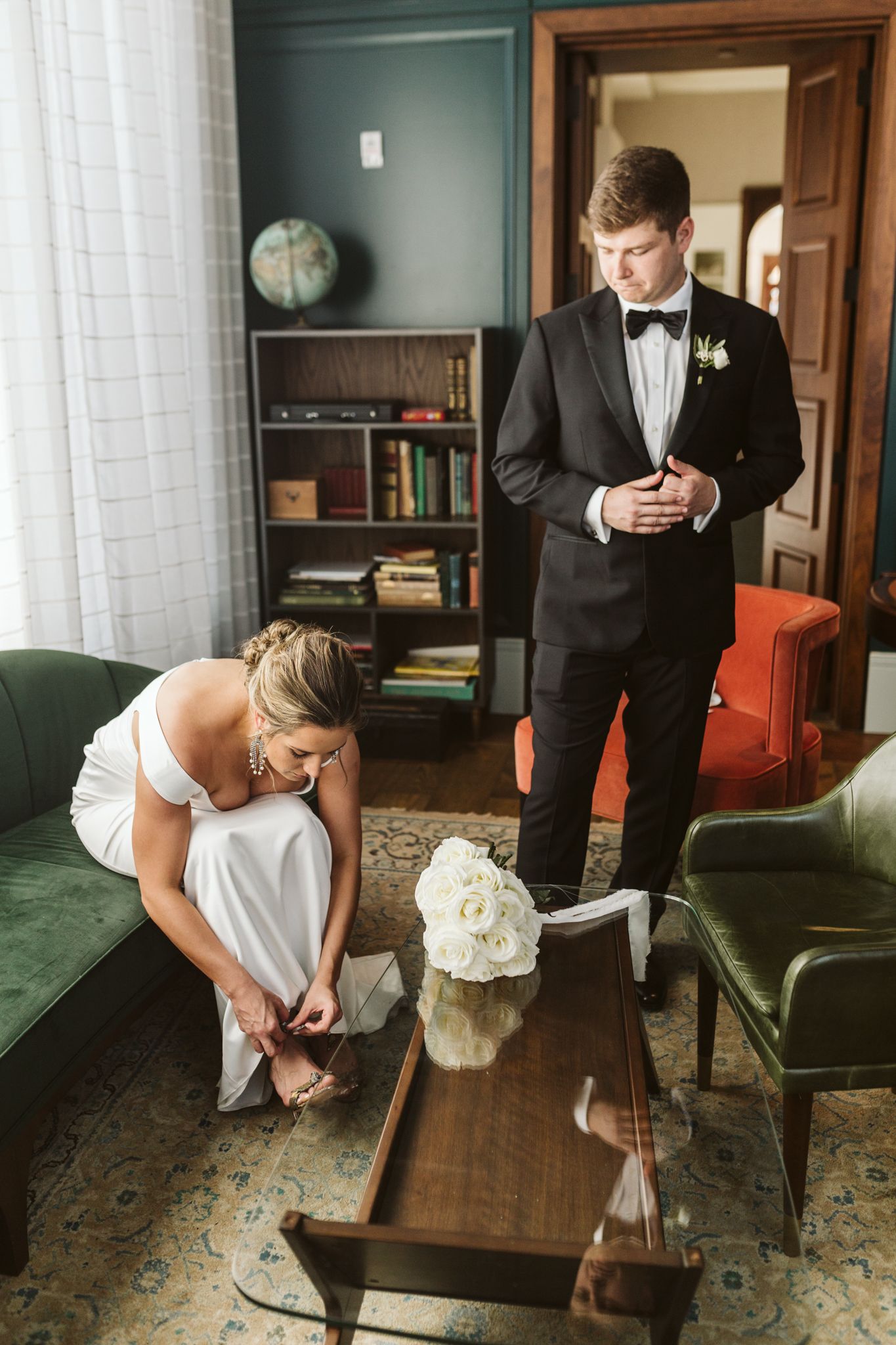 Bride adjusting her shoe in white gown while groom in black tuxedo buttons jacket in elegant vintage-styled room.