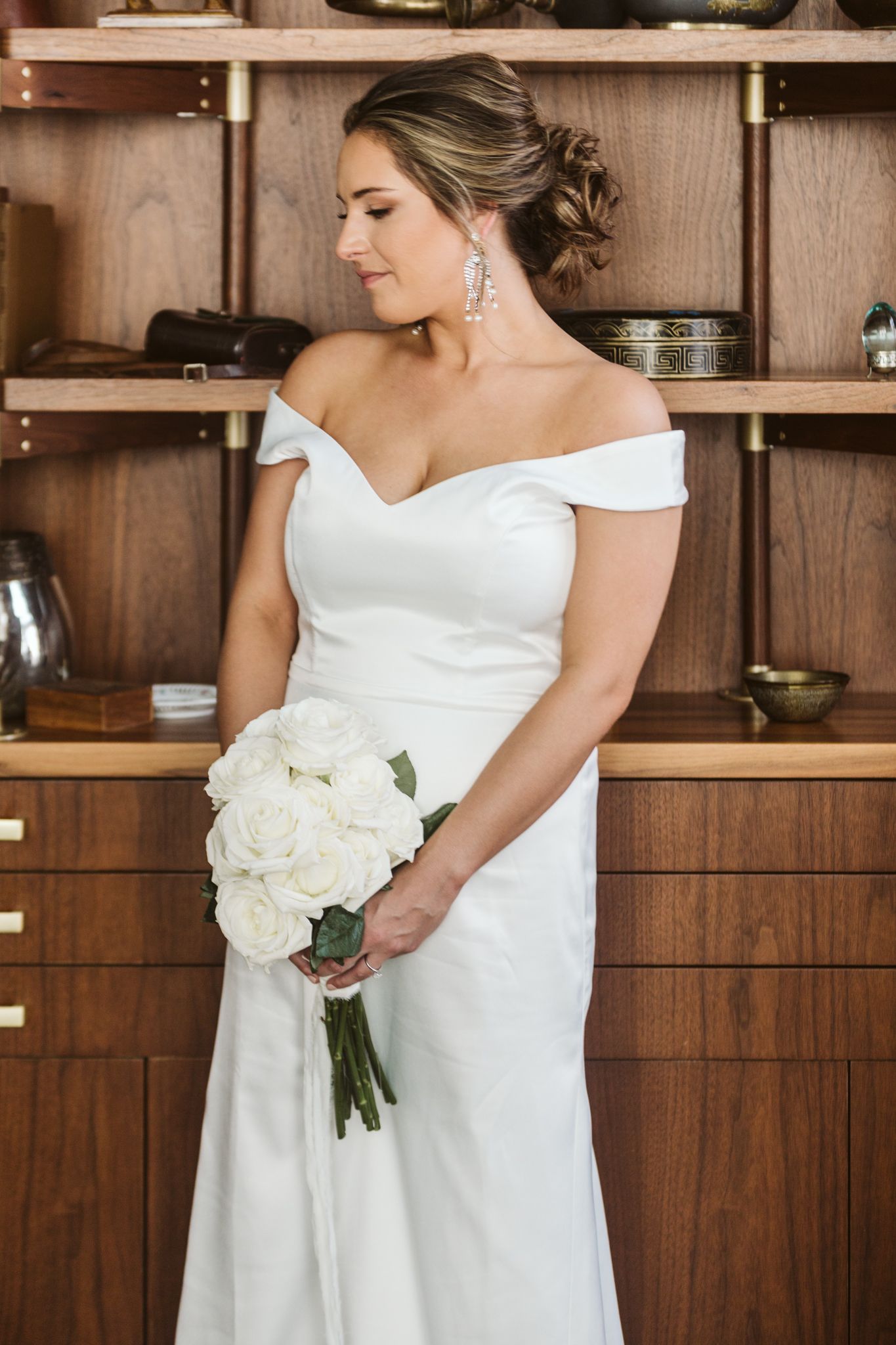 Bride in off-shoulder white gown holding white rose bouquet, standing before wooden shelving unit.