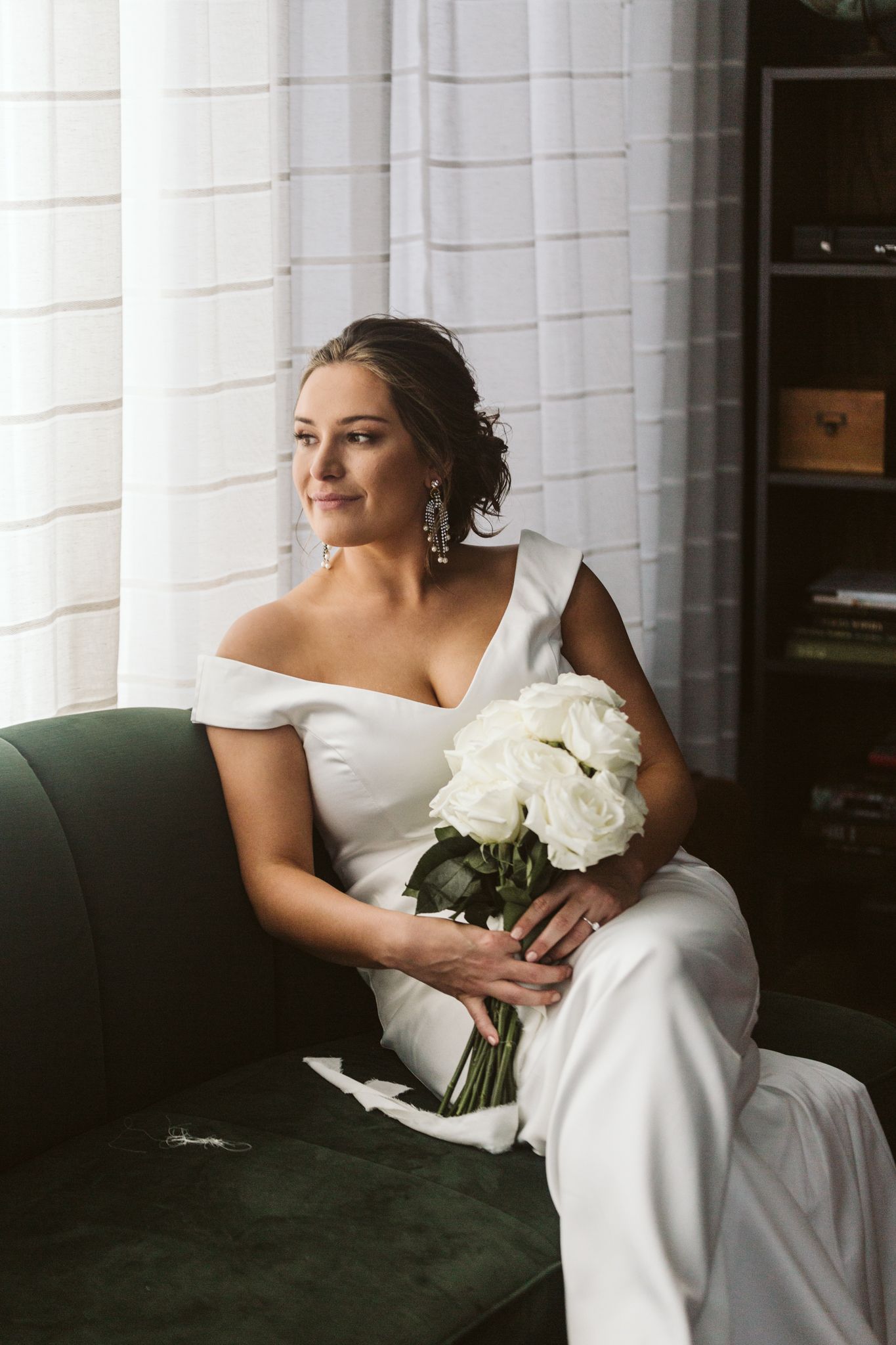 Bride in off-shoulder white gown holding white rose bouquet, seated on green velvet sofa near window.