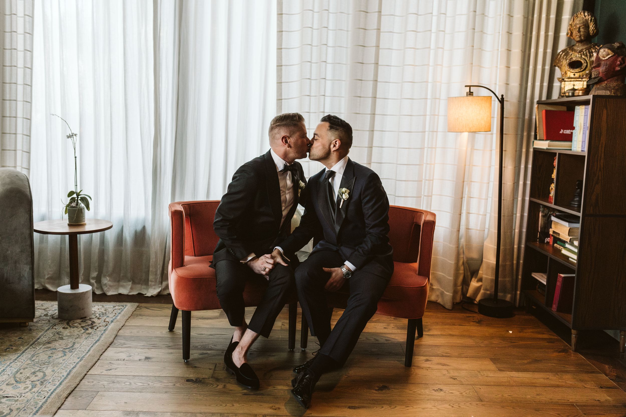Two grooms share a kiss while seated on a red chair in an elegantly decorated room on their wedding day.