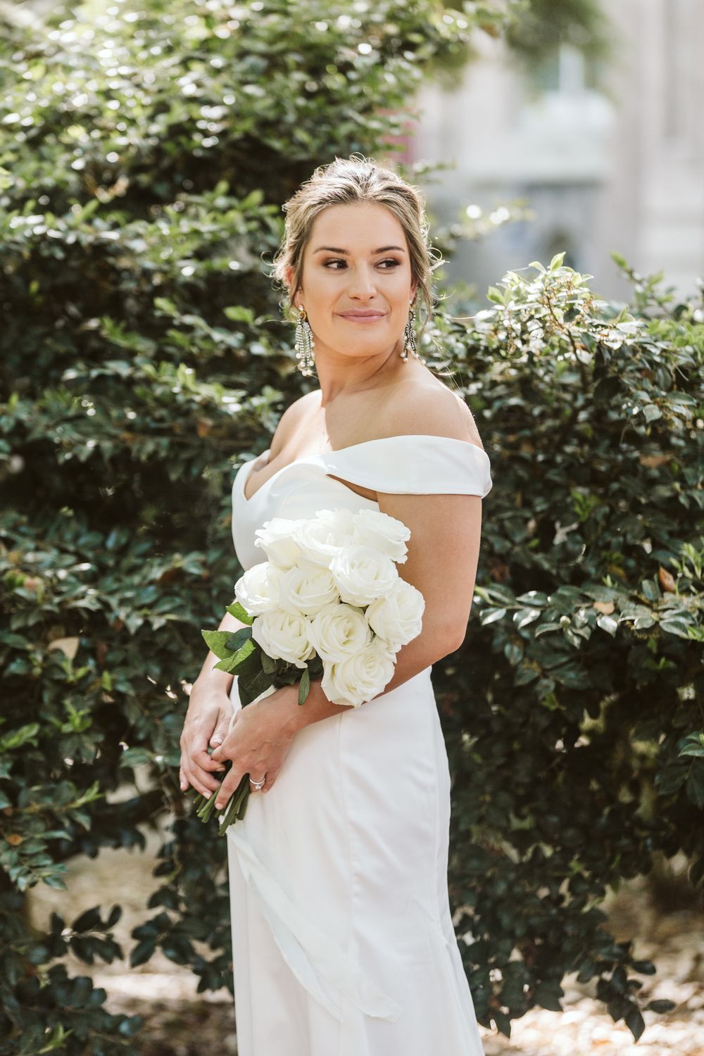 Bride in off-shoulder white gown holding white rose bouquet, posing outdoors near lush green hedges.